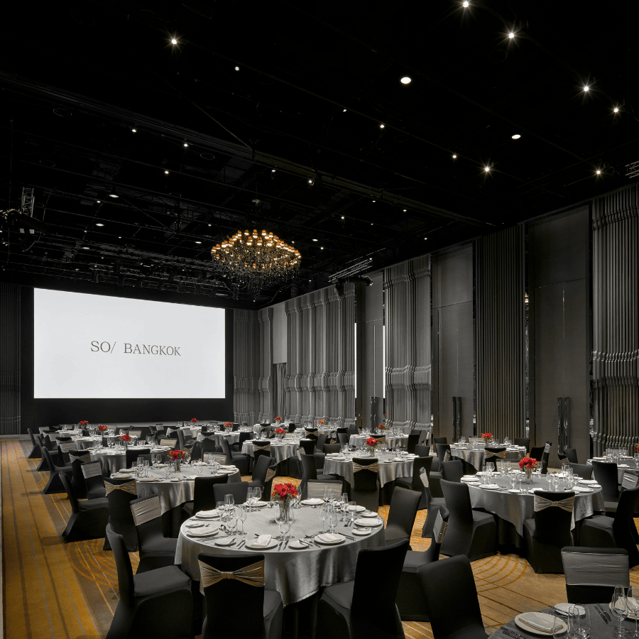 Several round fully set tables sit under a chandelier in a large ballroom with a large screen hung at the front.