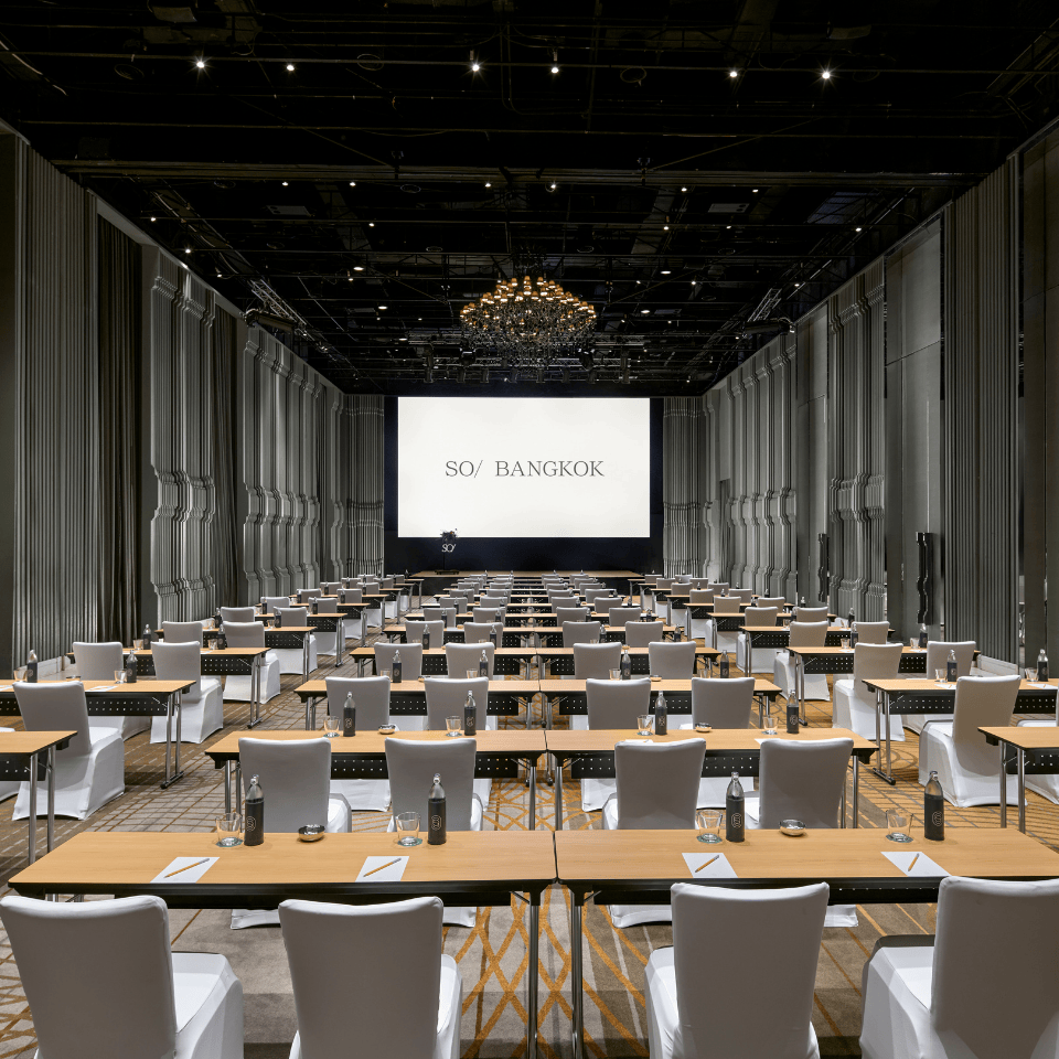 Conference room with rows of tables and white-covered chairs facing a large screen displaying a Bangkok cityscape and the words 'SO/ Bangkok'.