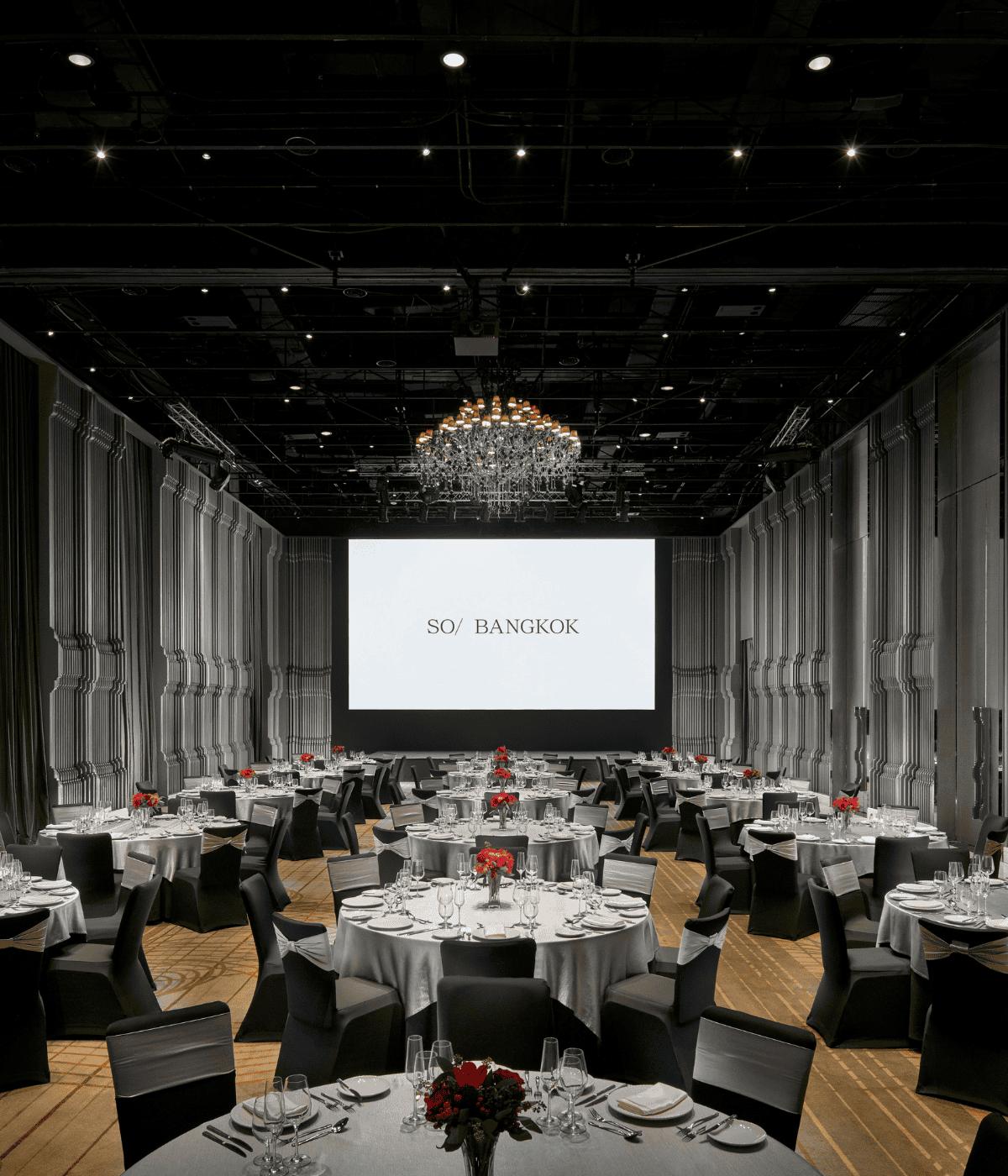 Elegant banquet setup in a design-led ballroom at SO/ Bangkok, featuring round tables, red floral centrepieces, and a grand chandelier beneath a large presentation screen.