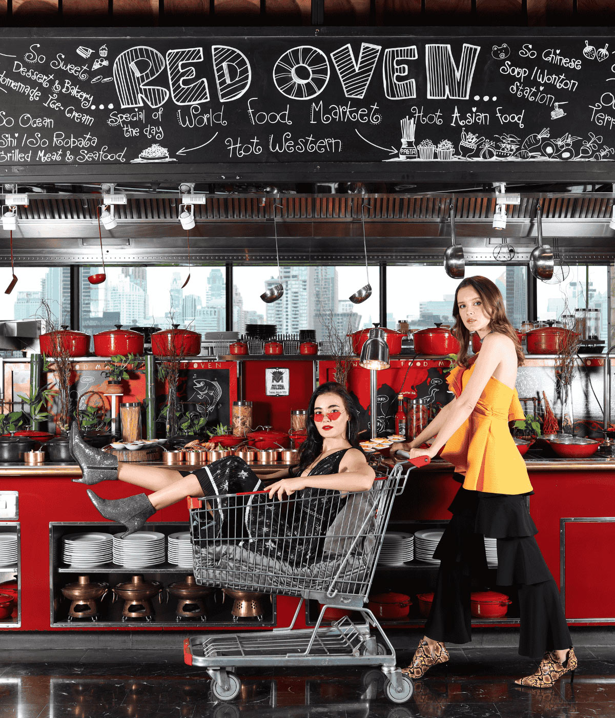Two stylish women posing playfully with a metal shopping cart in front of the Red Oven buffet counter, backed by red pots and a decorative chalkboard menu.
