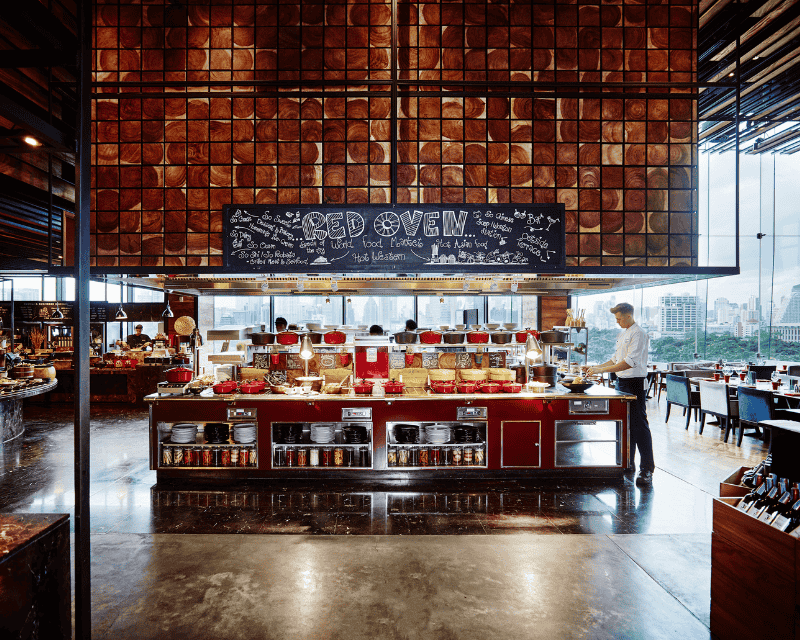 http://A%20professional%20chef%20preparing%20dishes%20behind%20the%20vibrant%20red%20counter%20of%20the%20Red%20Oven%20open%20kitchen,%20underneath%20a%20massive%20decorative%20wood-slice%20wall%20panel.
