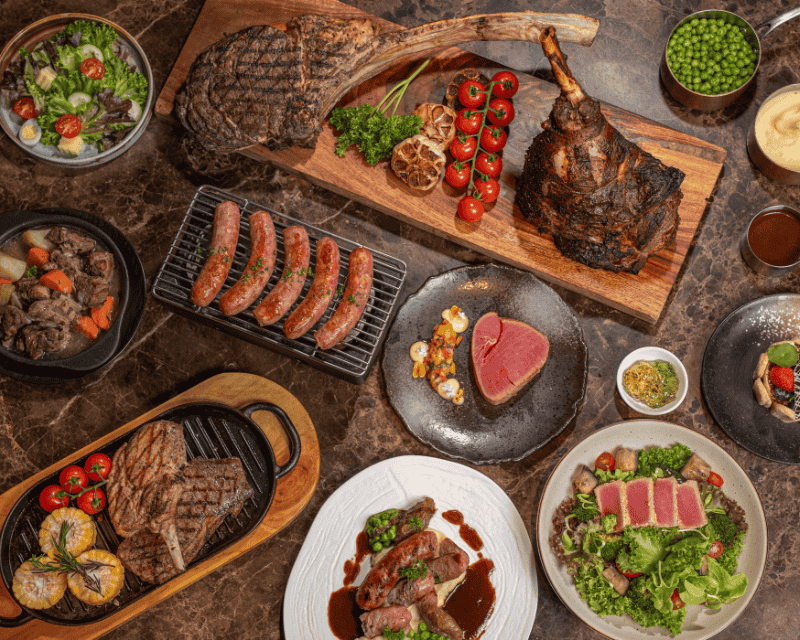 http://High-angle%20spread%20of%20a%20premium%20meat%20feast,%20featuring%20a%20massive%20Tomahawk%20steak,%20roasted%20lamb%20shank,%20grilled%20sausages,%20seared%20tuna%20steaks,%20and%20fresh%20green%20salads.