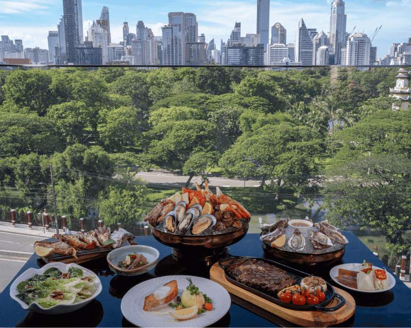 http://A%20premium%20selection%20of%20gourmet%20dishes%20including%20oysters,%20lobster,%20grilled%20steak,%20and%20salmon%20set%20on%20a%20dark%20table%20overlooking%20a%20green%20park%20and%20skyscrapers.