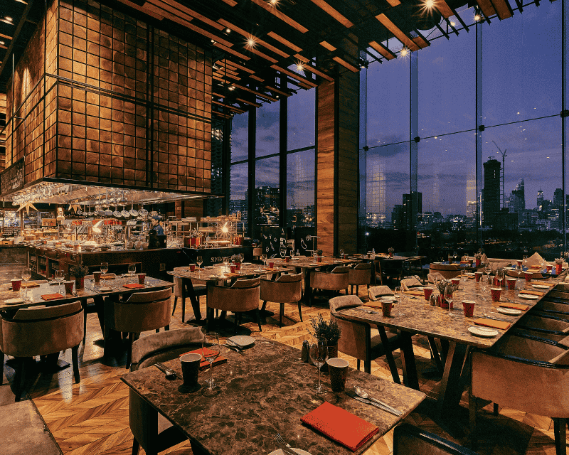 http://Warmly%20lit%20luxury%20restaurant%20at%20night%20with%20a%20stylish%20wooden%20ceiling%20structure,%20set%20tables,%20and%20a%20view%20through%20massive%20glass%20facades%20of%20the%20illuminated%20city%20skyline.