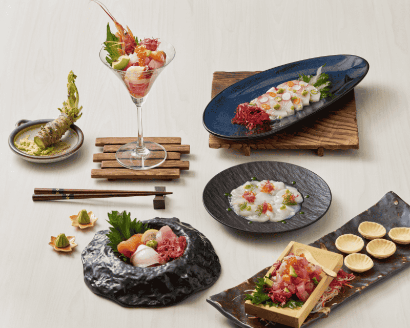 http://An%20artistic%20arrangement%20of%20Japanese%20seafood%20dishes%20on%20a%20light%20wooden%20table,%20including%20scallop%20carpaccio,%20tuna%20tartare%20in%20a%20martini%20glass,%20fresh%20sashimi%20in%20a%20stone%20bowl,%20and%20a%20whole%20wasabi%20root.