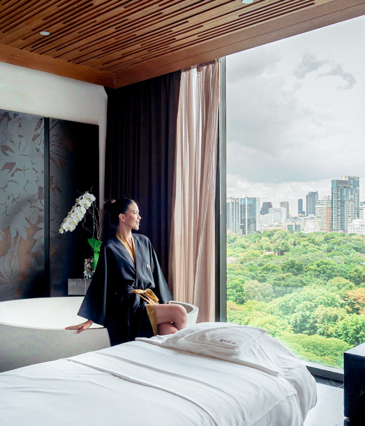 Close-up profile of a smiling woman in a black and gold robe sitting next to a white freestanding bathtub in a luxurious spa room with a city-view window. 