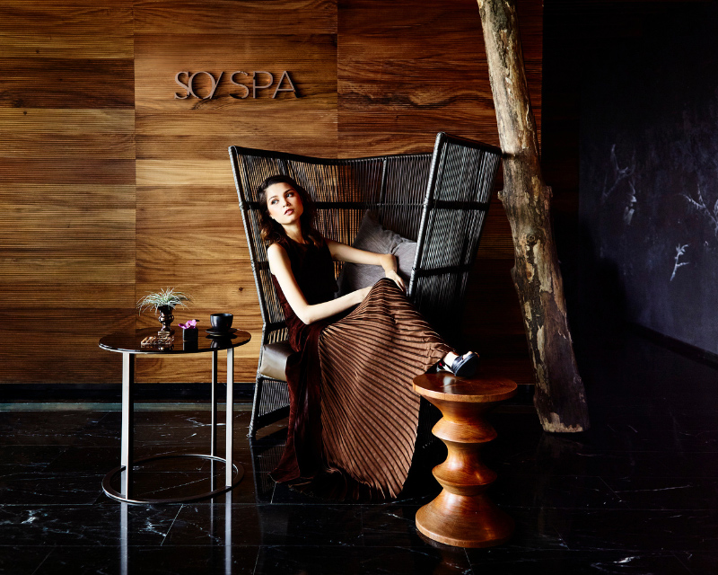 http://A%20young%20woman%20in%20a%20long%20brown%20pleated%20skirt%20sits%20elegantly%20in%20a%20high-backed%20dark%20woven%20armchair%20in%20front%20of%20a%20wood-paneled%20wall%20with%20the%20metallic%20SO/%20SPA%20logo.%20