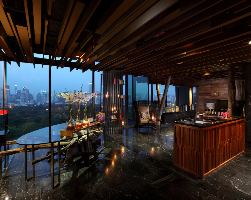 http://Spacious%20spa%20reception%20lounge%20at%20dusk%20featuring%20a%20wooden%20counter,%20a%20glass%20display%20table%20with%20white%20orchids,%20and%20a%20dramatic%20wall-sized%20window%20overlooking%20the%20city%20skyline%20under%20a%20deep%20blue%20twilight%20sky.
