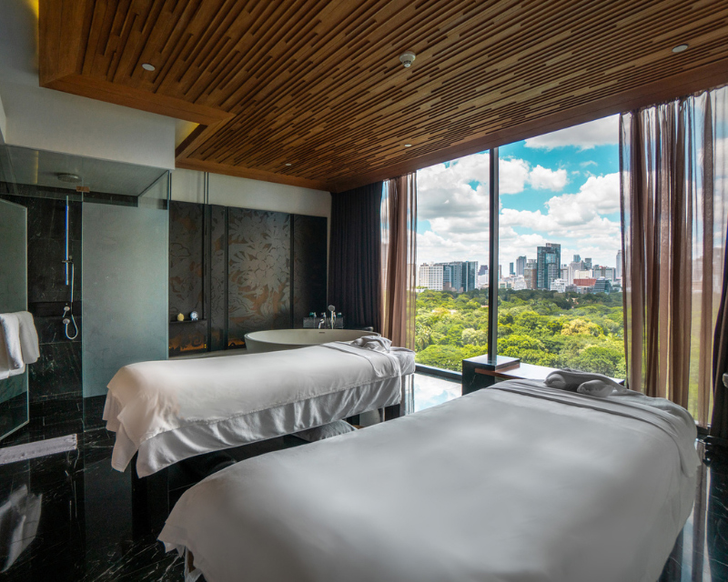 http://A%20luxury%20couple's%20spa%20treatment%20room%20with%20two%20massage%20beds,%20a%20deep%20soaking%20tub,%20and%20a%20floor-to-ceiling%20glass%20wall%20showcasing%20a%20panoramic%20view%20of%20Lumpini%20Park%20and%20the%20Bangkok%20skyline.