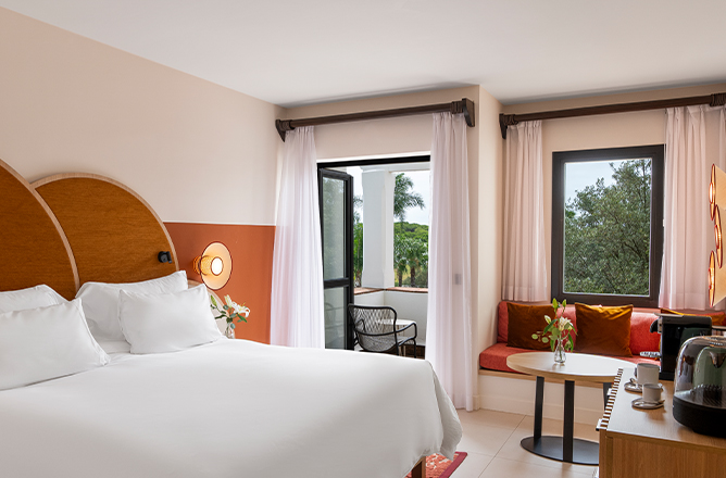 Habitación de hotel con cama king-size, cafetera y zona de estar. Una puerta balcón da al exterior con vistas a la vegetación.