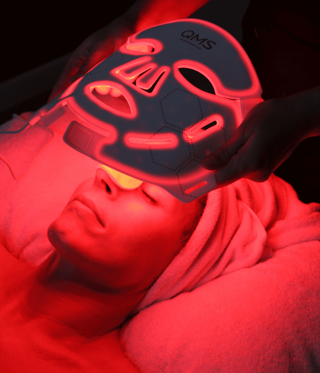 Person wearing a glowing red LED mask lying down with eyes closed, bathed in red light.