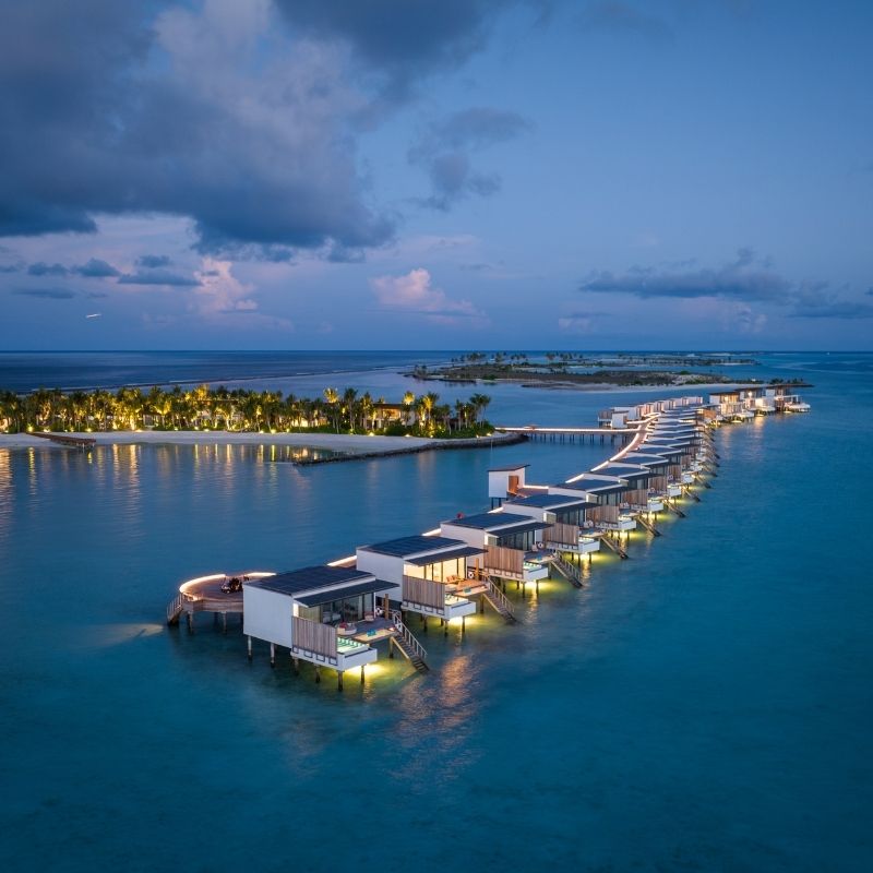 Aerial view of luxurious overwater villas illuminated at dusk, surrounded by turquoise waters and tropical greenery.