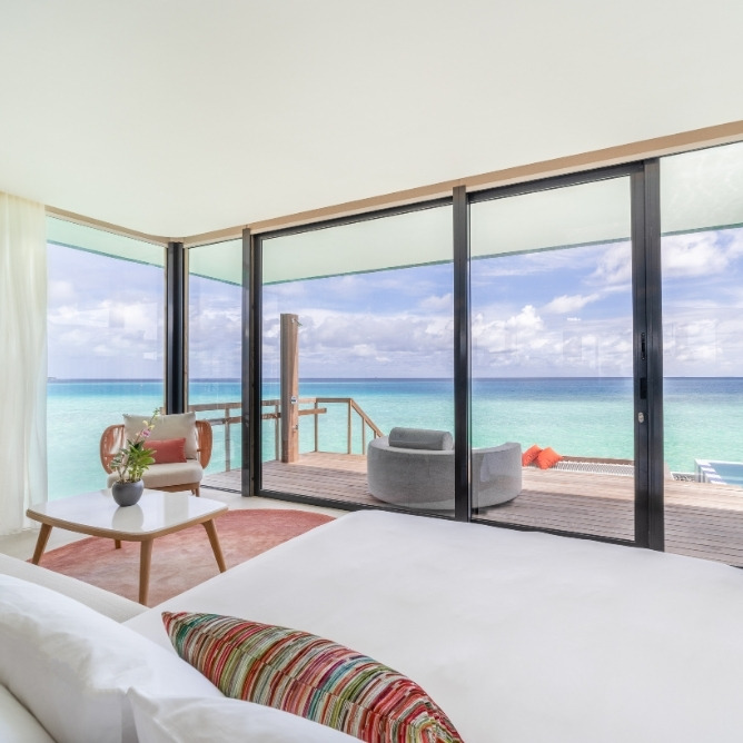 Modern bedroom with floor-to-ceiling windows showcasing a vibrant ocean view and a cozy outdoor deck area.