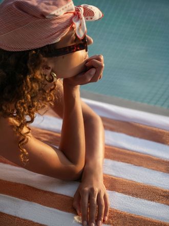 A person rests on a striped beach towel, wearing a stylish hat, with a serene pool in the background reflecting sunlight.