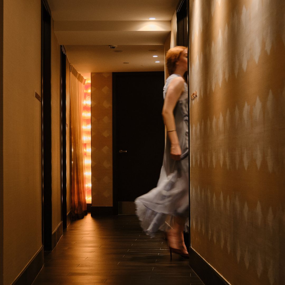 A woman in a flowing dress leans against a wall in a softly lit corridor, with warm lighting enhancing the elegant ambiance.