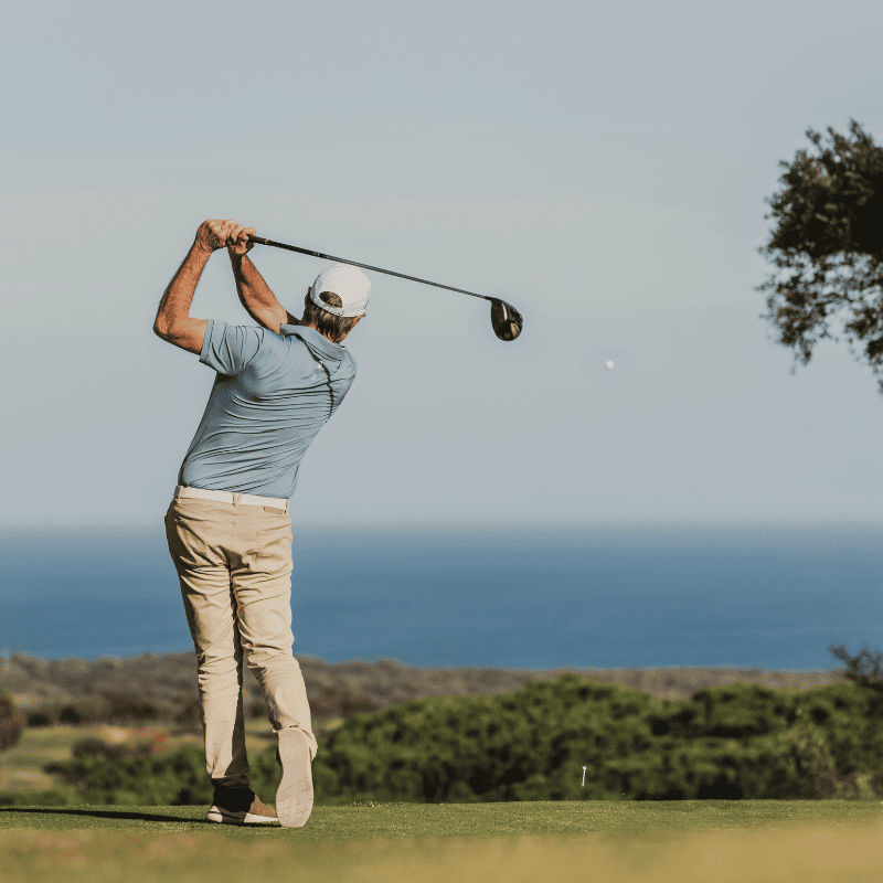 A man swings his golf club, with the ocean waves crashing in the background under a clear blue sky.