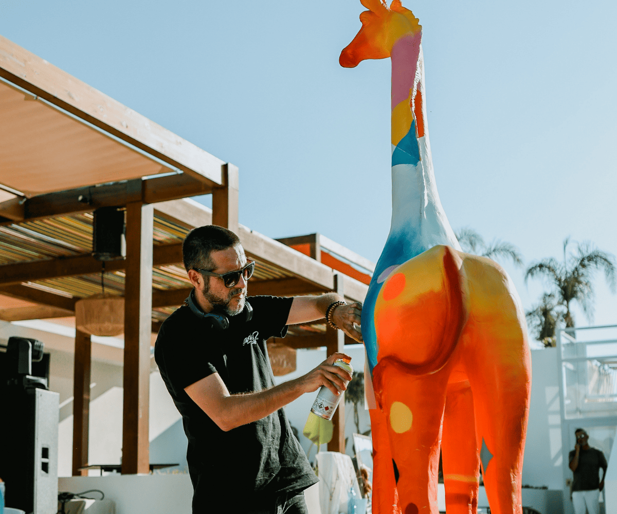 Un artiste peint à la bombe une sculpture de girafe aux couleurs vives et multicolores en plein air, entourée de palmiers et dans un décor moderne.