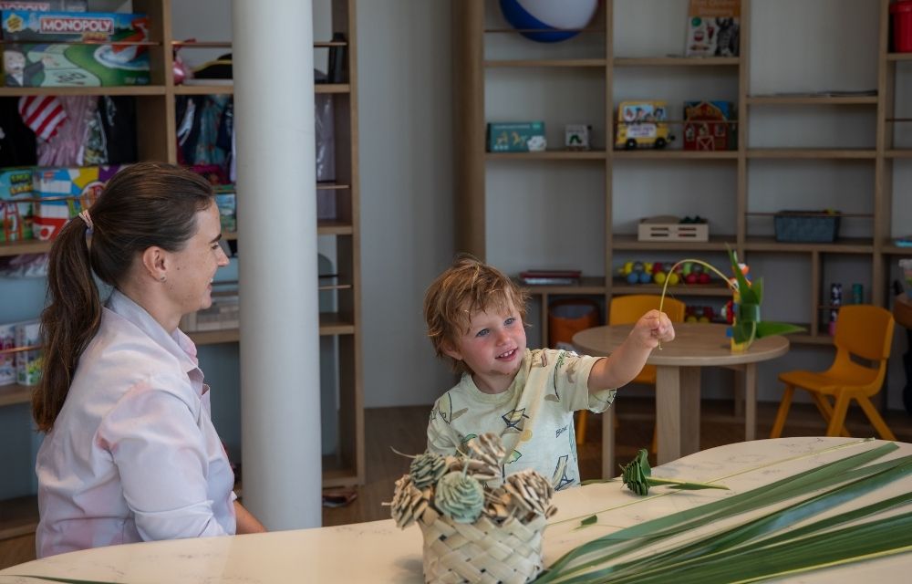 A woman and child are seated at a table at kids club, creating something from a palm leaf.