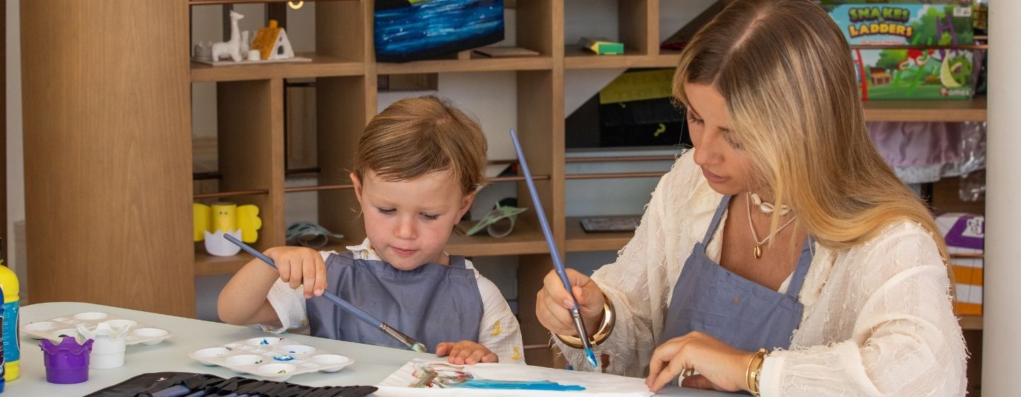 A woman and a child sit at a table with paintbrushes, creating art together at a kids club activity.