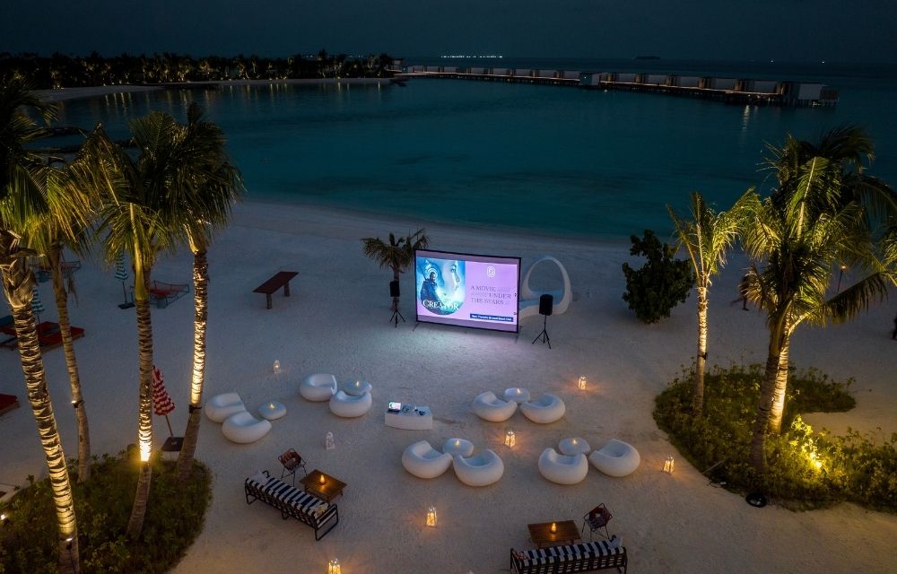 http://An%20outdoor%20movie%20screen%20illuminated%20at%20night,%20set%20against%20a%20beach%20backdrop%20with%20gentle%20waves.