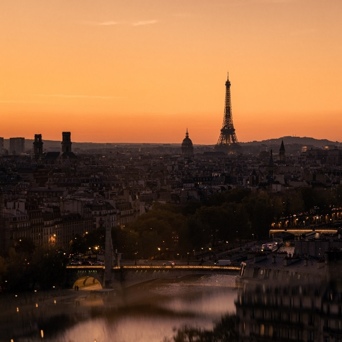A panoramic view of Paris at sunset, highlighting the illuminated Eiffel Tower against a vibrant orange sky.
