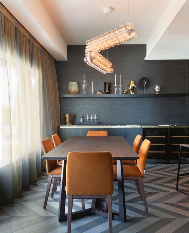 Modern dining area featuring a sleek table, orange chairs, elegant lighting, and dark decor with a herringbone floor pattern.