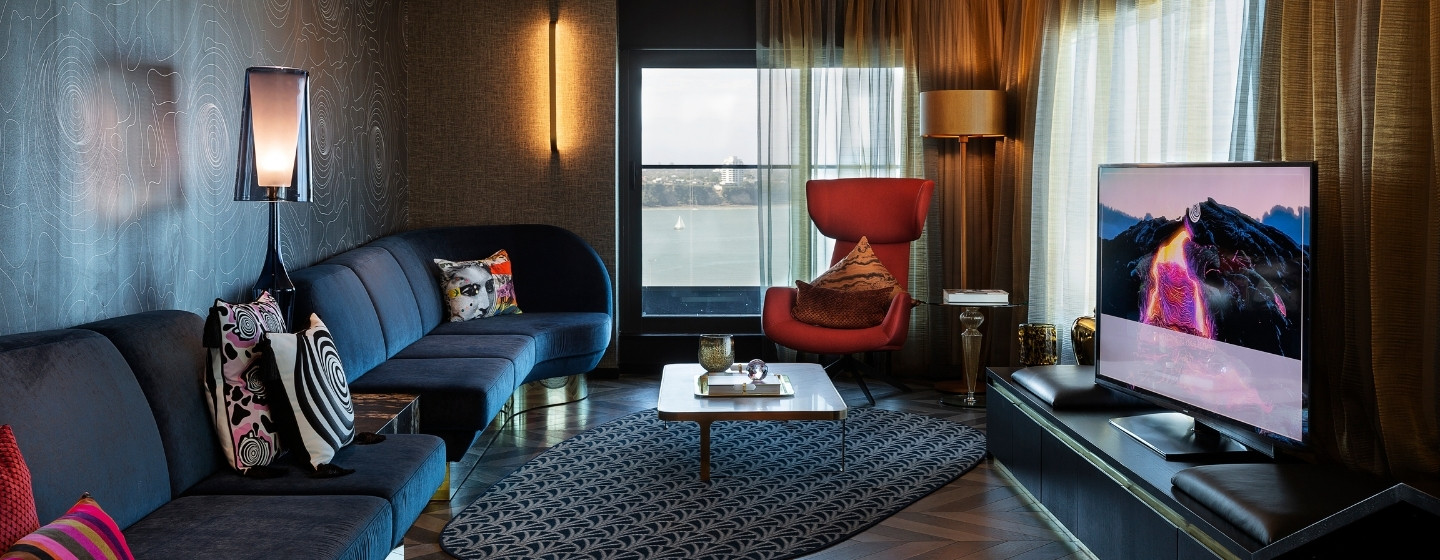 Modern living room with a curved blue sofa, red accent chair, stylish decor, large window, and a sleek television on a dark media unit.