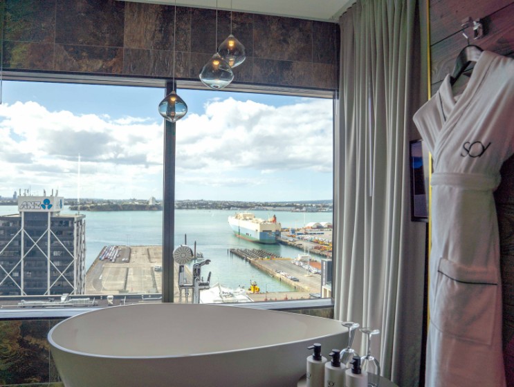 Spa-like bathroom with a freestanding tub, overlooking a harbor view through large windows, featuring soft curtains and elegant decor.