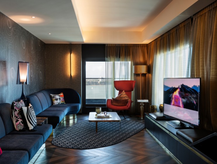 Modern living room with a curved blue sofa, red accent chair, stylish decor, large window, and a sleek television on a dark media unit.