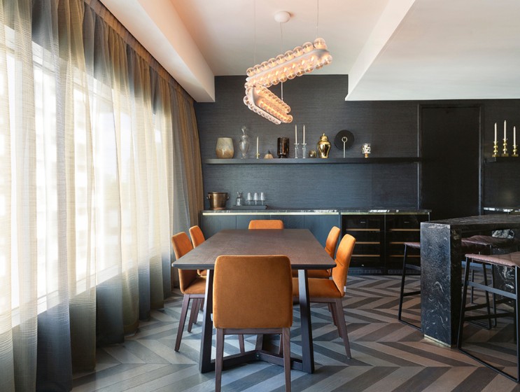 Modern dining area featuring a sleek table, orange chairs, elegant lighting, and dark decor with a herringbone floor pattern.