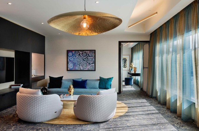http://Stylish%20living%20room%20featuring%20a%20gray%20sofa,%20plush%20chairs,%20golden%20lighting,%20and%20vibrant%20decor,%20set%20against%20sheer,%20blue%20curtains.