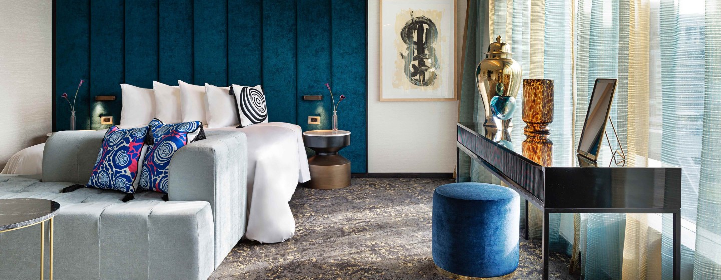A stylish hotel room featuring a teal accent wall, plush bedding, modern decor, and elegant light curtains.