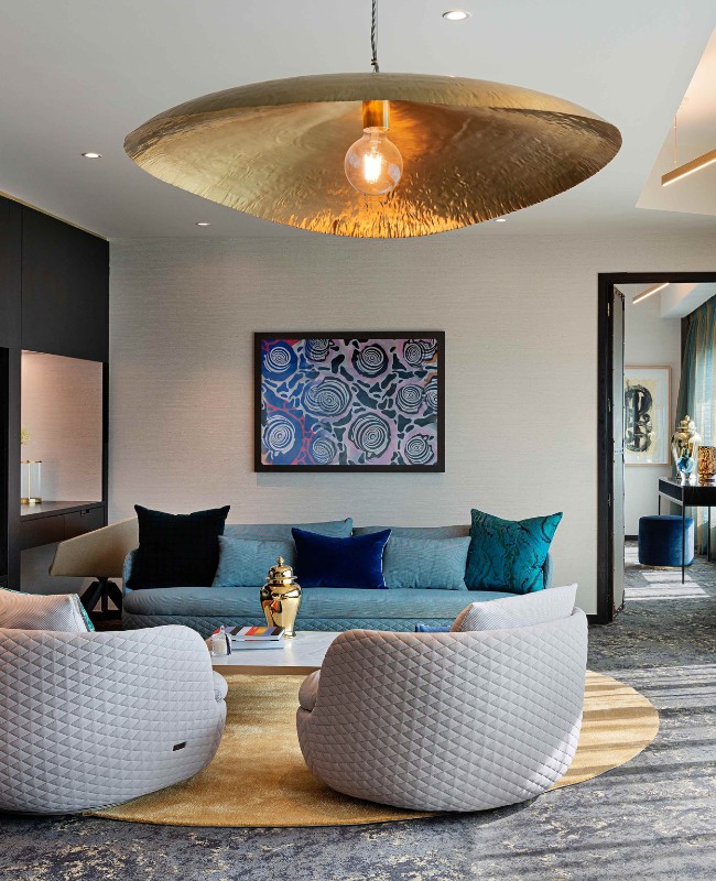 Stylish living room featuring a gold pendant light, modern art, a blue sofa, and quilted armchairs on a round rug.