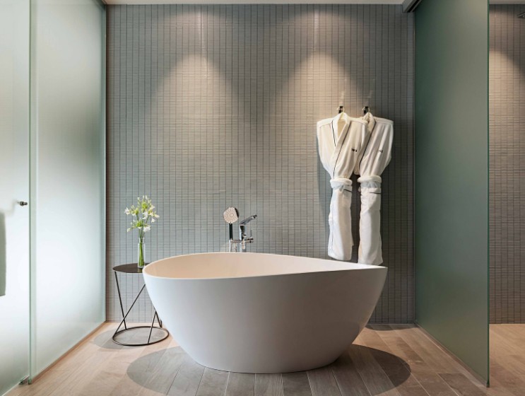 A modern bathroom featuring a sleek white freestanding tub, green wall, glass shower, stylish stool, and hanging bathrobes.