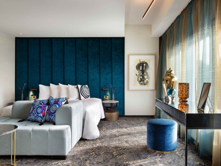 A stylish hotel room featuring a teal accent wall, plush bedding, modern decor, and elegant light curtains.
