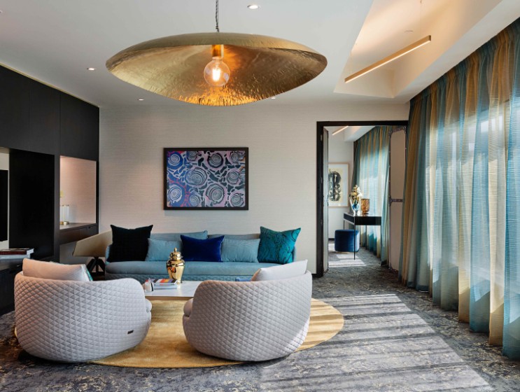 Stylish living room featuring a gray sofa, plush chairs, golden lighting, and vibrant decor, set against sheer, blue curtains.