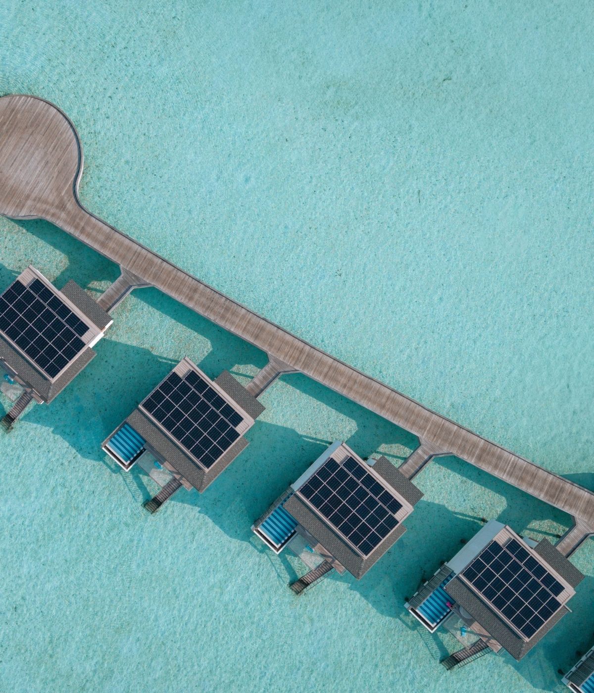 Aerial view of overwater villas with solar panels, connected by a wooden walkway over crystal-clear turquoise waters.