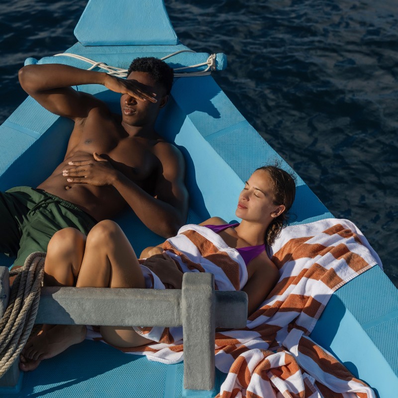 A couple relaxes on a blue boat, wrapped in striped towels, enjoying the sun and sea under clear skies.