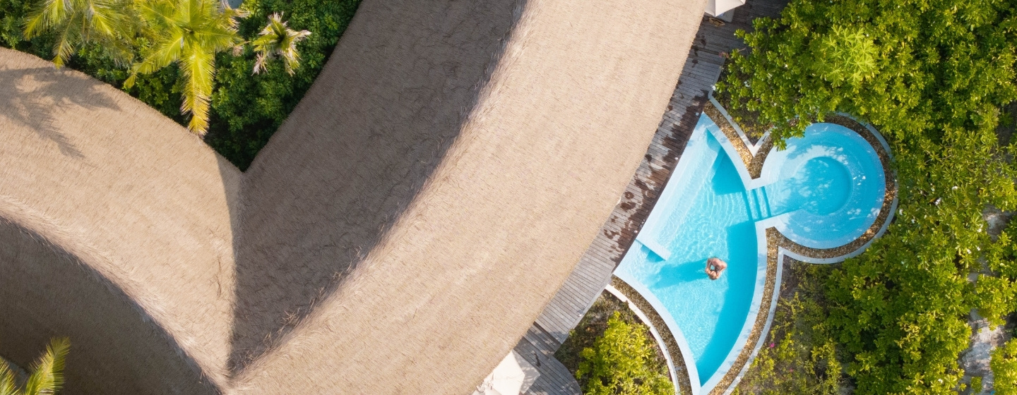 Aerial view of a luxurious pool, surrounded by greenery, with a person relaxing in the water beneath a thatched roof.