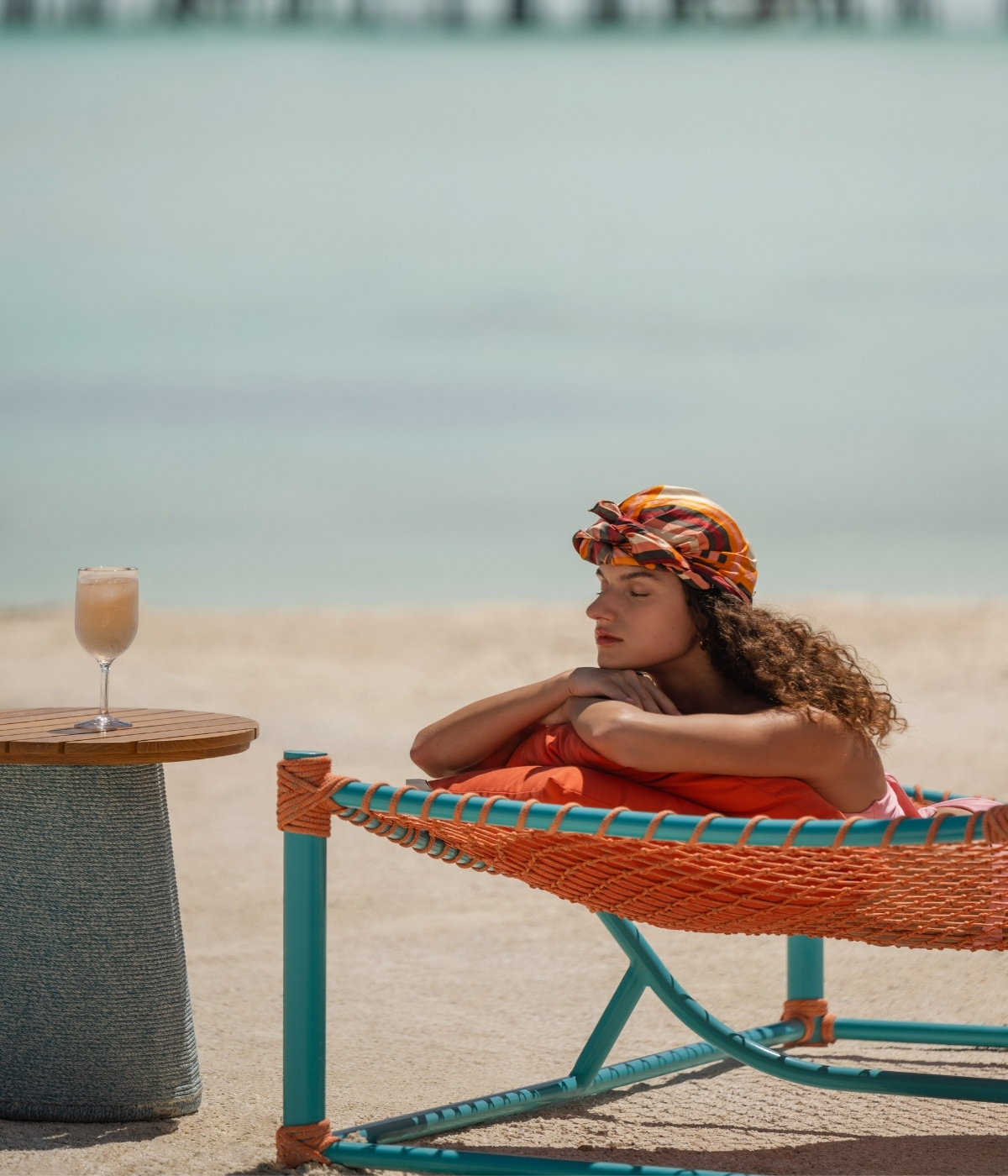 A woman relaxes on a beach hammock, wrapped in a vibrant orange blanket, beside a wooden table with a glass of sparkling drink.