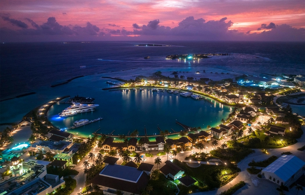 Aerial view of a luxurious resort at twilight, featuring illuminated villas, yachts in the marina, and vibrant sunset colors over the ocean.