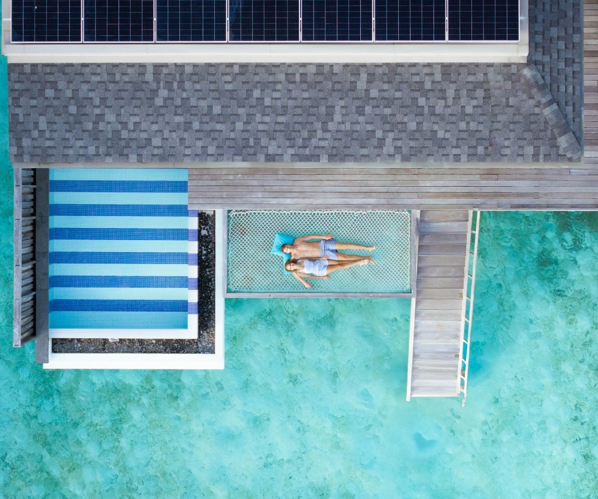 Aerial view of a couple relaxing on a hammock by a turquoise sea, with a pool and solar panels in the background.