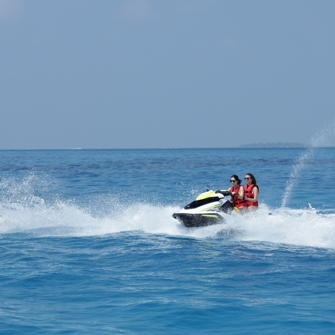 Two women in life jackets ride a jet ski across vibrant blue waters, creating splashes as they enjoy a sunny day on the ocean.