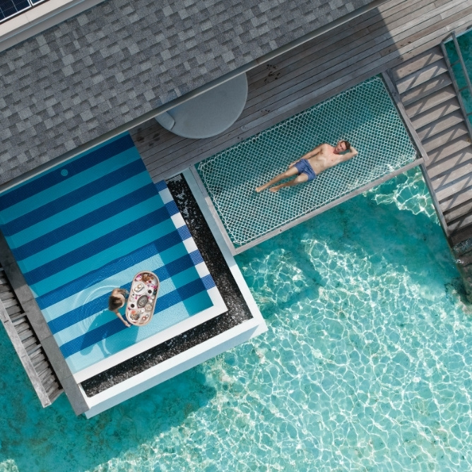 Aerial view of a resort pool with a person on a vibrant floatie and another lounging on a deck, surrounded by clear turquoise water.