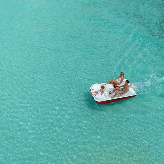 Three people enjoy a pedal boat ride on crystal-clear turquoise waters under the sun, surrounded by a serene tropical setting.