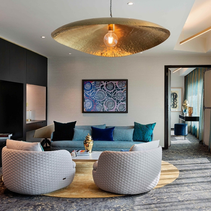 Stylish living room featuring a gold pendant light, modern art, a blue sofa, and quilted armchairs on a round rug.