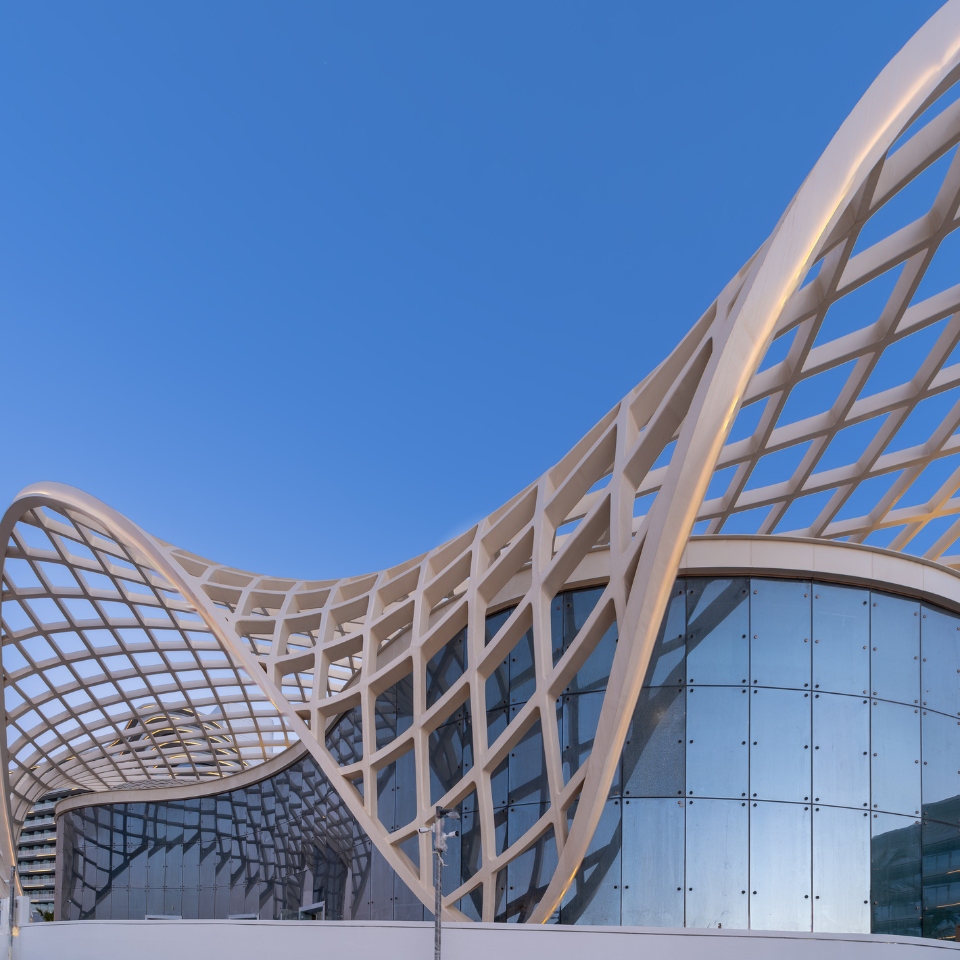 Modern architectural structure with a curved lattice roof above glass walls, set against a clear blue sky.