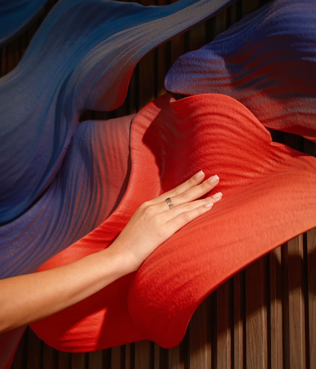 A hand with a ring gently touches flowing, textured fabric in red and blue hues. The motion creates a wavy, dynamic visual against a wooden backdrop.