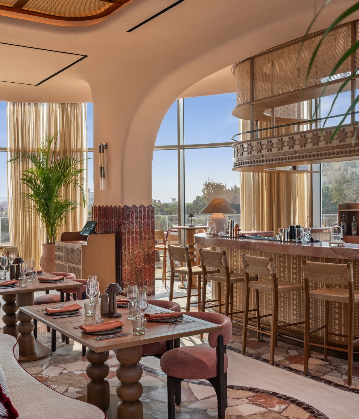 Stylish restaurant interior featuring a wooden bar, green chairs, elegant tables, and warm lighting with large windows.