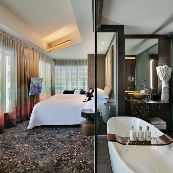Stylish hotel room featuring a bed, modern decor, a glass-enclosed bathroom with a bathtub, and panoramic windows with sheer curtains.